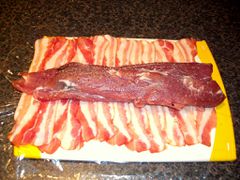 Das gefüllte und gepfefferte Schweinefilet unter Zuhilfenahme von Frischhaltefolie mit Bacon ummanteln.